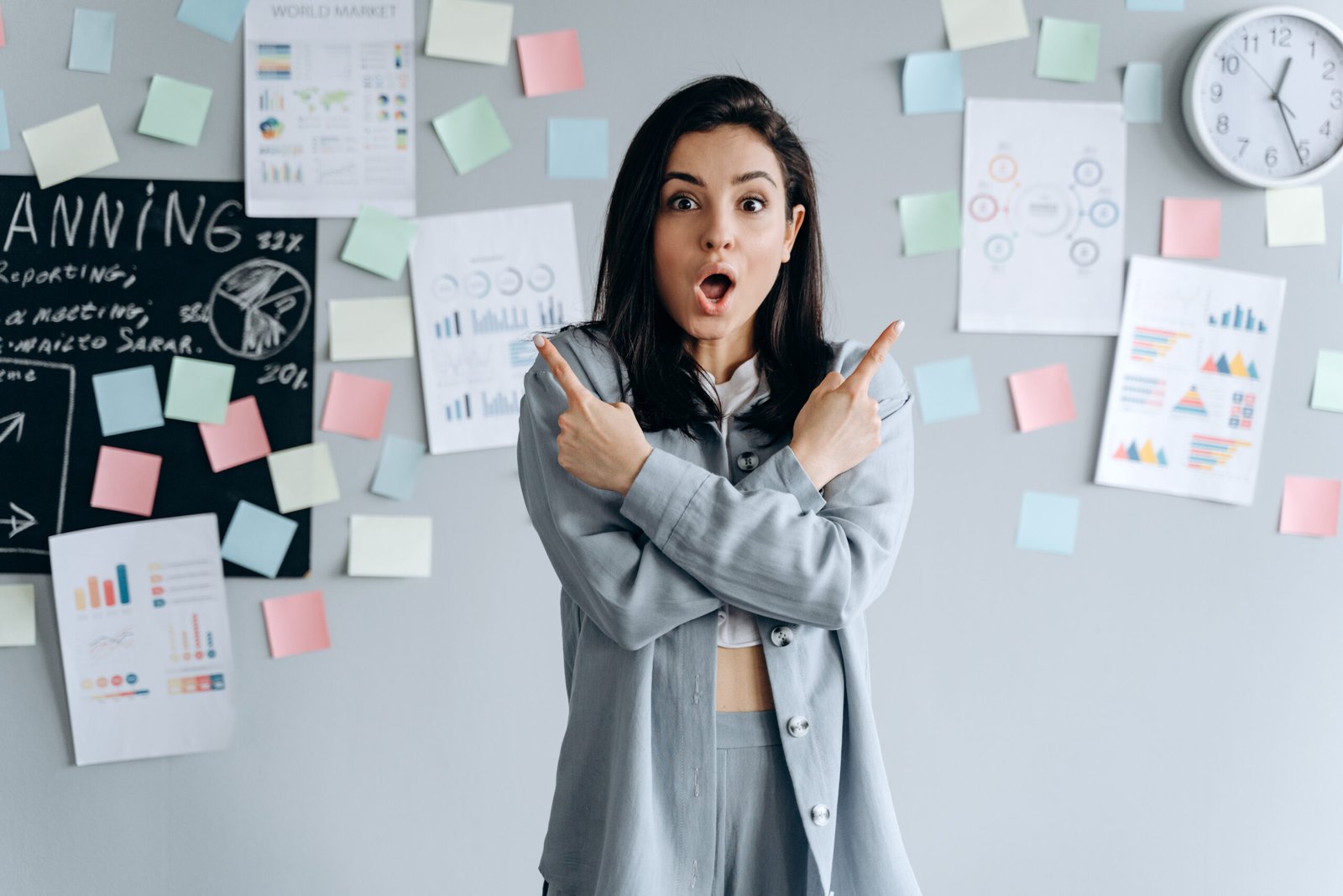 Beautiful, business girl on a gray work background shows index fingers up, arms crossed. Cute young woman in the office.
